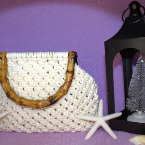May include: Cream-colored macrame handbag with bamboo handles. A black lantern with a small Christmas tree inside is to the right. Two white starfish are in front of the bag, with a purple background.