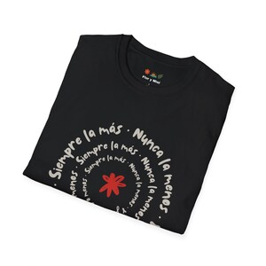 May include: Black t-shirt with a circular design in white script, featuring the words "Siempre la más. Nunca la menos." A red star is at the center of the design. The shirt also has the text "Flor y Miel" near the collar.