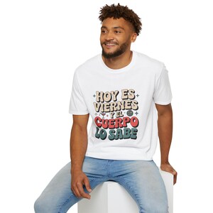 May include: White t-shirt featuring the Spanish text "Hoy es Viernes y el Cuerpo lo Sabe" in a retro style. The text is in beige, red, and green. The person is wearing blue jeans.