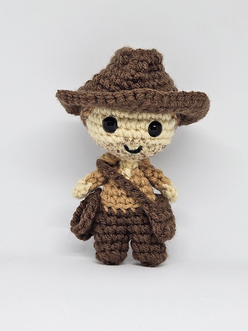 May include: A crocheted toy figure of a person wearing a brown cowboy hat and brown and tan clothing. The toy has a smiling face with black eyes and a brown nose.