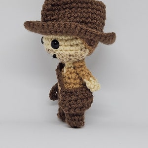 May include: A crocheted brown and cream cowboy doll wearing a brown hat. The doll is standing on a white background.