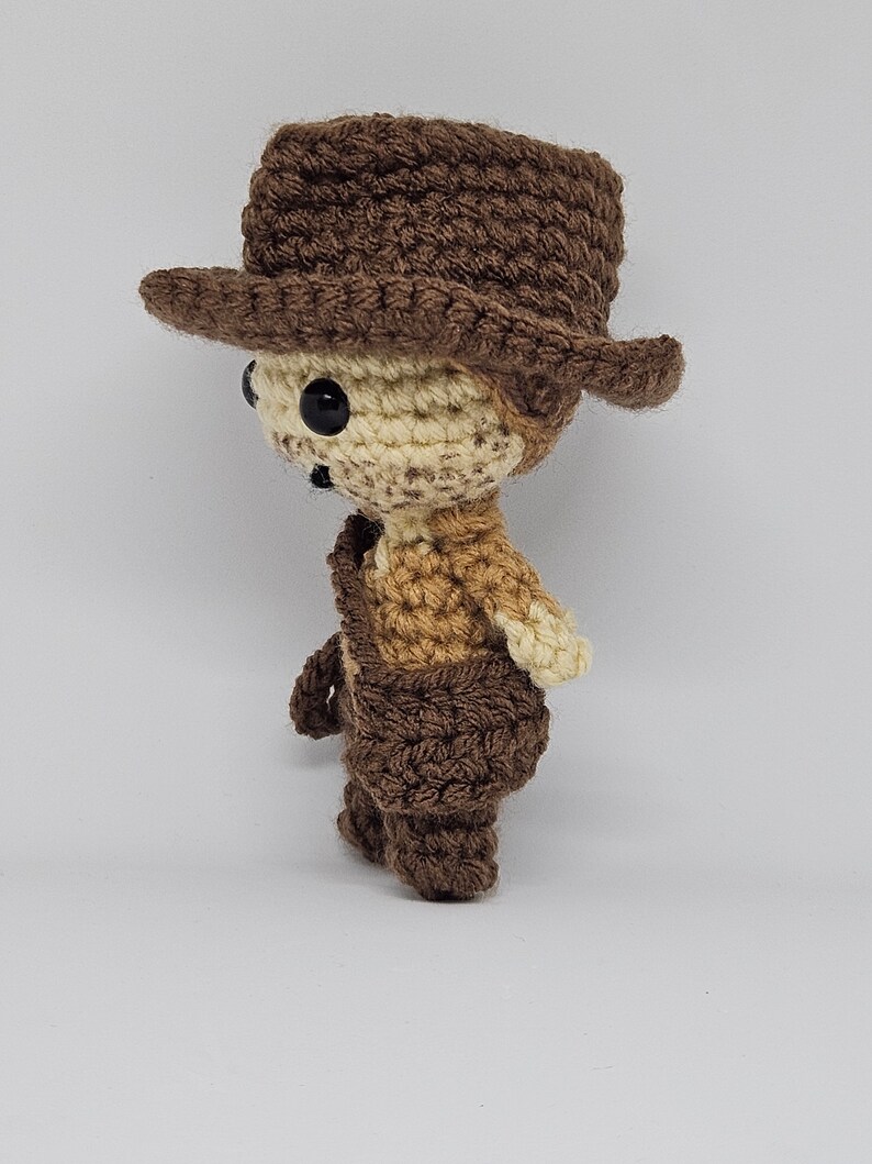 May include: A crocheted brown and cream cowboy doll wearing a brown hat. The doll is standing on a white background.
