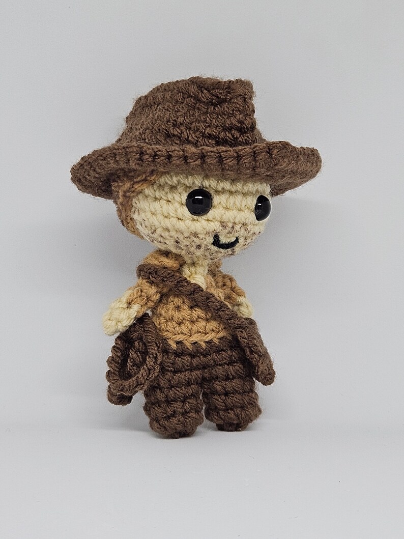 May include: A crocheted toy figure wearing a brown cowboy hat and brown and beige clothing. The toy has a smiling face with black eyes.