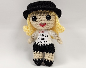 Peut inclure: Une poupée en crochet portant un chapeau noir, une chemise blanche avec le texte "A LOT GOING ON AT THE MOMENT" et un short rayé noir et blanc.