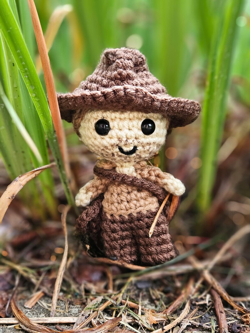May include: A crocheted amigurumi doll wearing a brown hat with a wide brim. The doll has a smiling face with brown eyes and is standing in a bed of green grass and brown leaves.