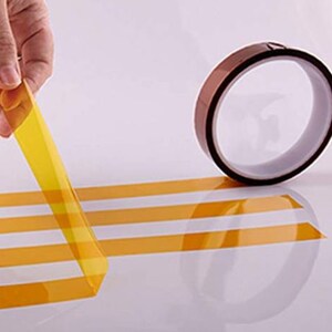 Puede incluir: Un rollo de cinta resistente al calor de color amarillo con un respaldo marr&oacute;n. La cinta se est&aacute; desenrollando y est&aacute; sobre una superficie blanca.