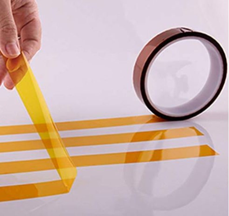 Puede incluir: Un rollo de cinta resistente al calor de color amarillo con un respaldo marr&oacute;n. La cinta se est&aacute; desenrollando y est&aacute; sobre una superficie blanca.