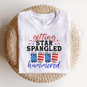 May include: White t-shirt with a red, white, and blue design. The design features four cans of beer with the American flag printed on them. The text "getting star spangled hammered" is printed on the shirt.