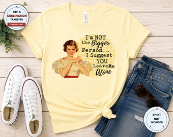 May include: A pale yellow short-sleeved t-shirt featuring a vintage-style illustration of a woman and the text, I'm NOT the Bigger Person... I Suggest YOU Leave Me Alone, printed over a light yellow checkered circle background.