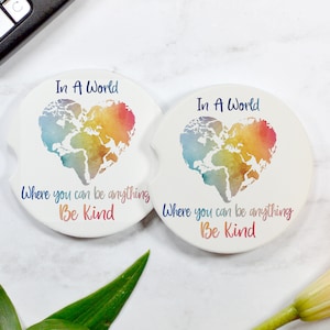 May include: Two round white coasters with a watercolor world map in the shape of a heart. The text "In A World Where You Can Be Anything Be Kind" is printed on each coaster.