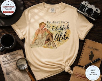 May include: A beige t-shirt featuring a graphic of a vintage-style woman in a dress and stockings, with the text "I'm Just Here to Establish an Alibi" in black script and print lettering, set against a soft yellow and brown watercolor-style background.