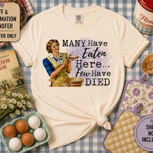May include: A cream-colored short-sleeve t-shirt featuring a vintage-style illustration of a person in an apron holding a bowl, with the text "MANY Have Eaten Here... Few Have DIED" printed in black, set against a light purple polka dot background.
