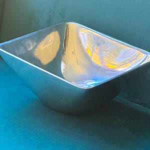 May include: A square-shaped bowl with a polished, reflective silver-toned metallic finish. The bowl features smooth, gently curved sides that taper toward a smaller base, creating a wide, open top with rounded corners and a clean, seamless edge.