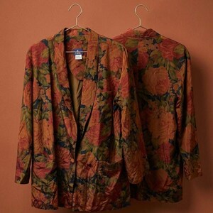 Puede incluir: Dos blazers con estampado floral colgando de perchas doradas. Los blazers presentan un patrón de grandes rosas en tonos naranja, rojo y verde sobre un fondo azul oscuro. Los blazers tienen dos bolsillos delanteros y cierre de botón.