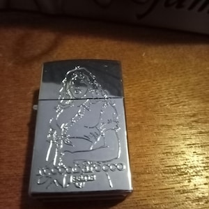 Puede incluir: Un encendedor plateado con un diseño en relieve de una mujer. El encendedor rectangular tiene una tapa con bisagras. El diseño incluye el texto "Bonne Nuit Rocco" y "Paris".