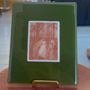 May include: A rectangular olive green glass plaque featuring a centered white inset panel. The white panel displays a sepia-toned photographic image of a couple in wedding attire walking outdoors among trees. The plaque rests on a gold metal stand.