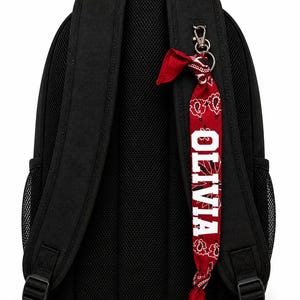 May include: A black backpack featuring padded shoulder straps and side mesh pockets. A red bandana with a white paisley pattern is attached to the right strap using a silver metal clip. The name OLIVIA is printed in white block letters on the bandana.