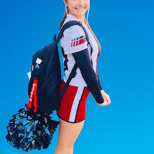 May include: A cheerleader in a white, red, and black uniform with a backpack and a black pom-pom. The uniform includes a white top with red and black accents, a red skirt with a white stripe, and black sleeves. The backpack has a red tag that says "OLIVIA".