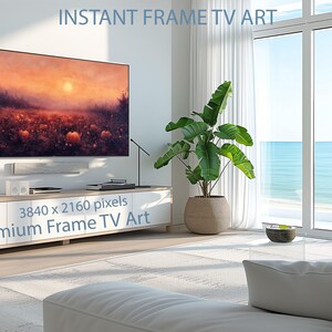 May include: A large flat-screen TV displays a sunset landscape with pumpkins. The TV is mounted on a white wall above a modern console. The room features a large window with a view of the ocean, a potted plant, and a white sofa. Text on the screen reads "INSTANT FRAME TV ART".