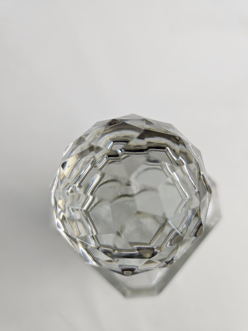 K&ouml;nnte beinhalten: Ein klares, facettiertes Glasobjekt mit geometrischem Design. Das Objekt hat eine abgerundete Oberseite und eine flache, sechseckige Basis. Die Facetten fangen das Licht ein und erzeugen einen funkelnden Effekt. Es ist wahrscheinlich ein dekorativer Gegenstand oder ein Beh&auml;lter.