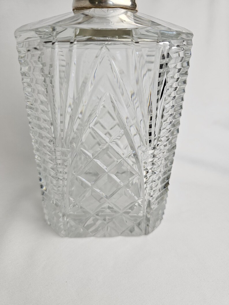 K&ouml;nnte beinhalten: Eine klare, rechteckige Glasdekanter mit einem silberfarbenen Verschluss. Der Dekanter hat ein Diamanten- und vertikales Linienmuster. Das Glas ist dick und hat ein facettiertes Design, das einen funkelnden Effekt erzeugt. Der Dekanter ist leer.