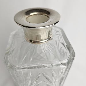 K&ouml;nnte beinhalten: Ein klarer, facettierter Glasparf&uuml;mflakon mit einem silberfarbenen Metallkragen. Die Flasche hat ein geometrisches Design und eine breite, kreisf&ouml;rmige &Ouml;ffnung. Das Glas ist transparent.