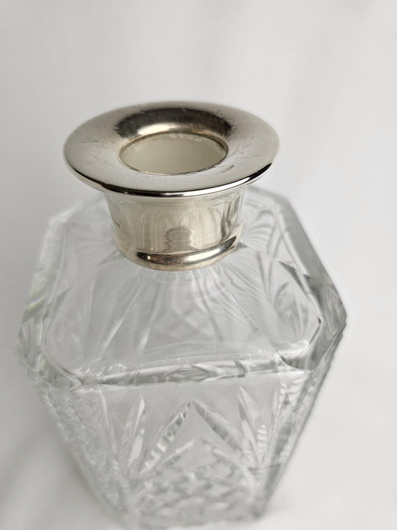 K&ouml;nnte beinhalten: Ein klarer, facettierter Glasparf&uuml;mflakon mit einem silberfarbenen Metallkragen. Die Flasche hat ein geometrisches Design und eine breite, kreisf&ouml;rmige &Ouml;ffnung. Das Glas ist transparent.