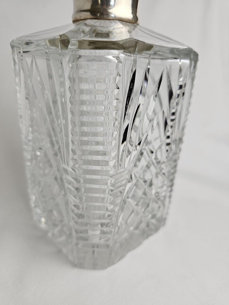 K&ouml;nnte beinhalten: Eine klare, facettierte Glasparf&uuml;mflasche mit silberfarbenem Deckel. Die Flasche hat ein geometrisches Design mit abgewinkelten Schnitten und eine rechteckige Form. Der Deckel ist aus glattem, metallischem Material. Die Flasche ist leer und steht vor wei&szlig;em Hintergrund.