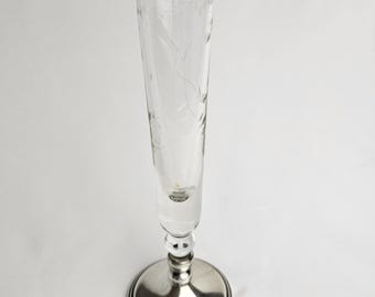 May include: A clear, etched glass champagne flute with a silver-colored base. The glass features a delicate floral design. The base is round and has a detailed, layered design. The flute is empty and stands upright.