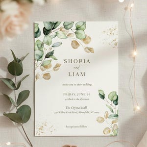 Puede incluir: Invitación de boda con los nombres "Shopia y Liam" y detalles. La invitación presenta hojas de eucalipto en acuarela y detalles dorados. El texto incluye la fecha, hora y lugar: The Crystal Hall, Bloomfield, NY.