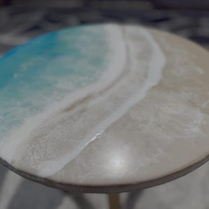May include: Round table with a resin top depicting a beach scene. The design features shades of blue, white, and beige, resembling ocean waves and sand. The table has a wooden edge and a gold-colored base.