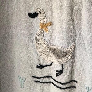 May include: A light blue textured towel featuring a central embroidered white goose with a yellow neck ribbon, black beak, and black feet. The design includes a small yellow butterfly, a cluster of pink and blue flowers, and black wavy water lines.