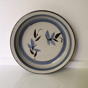 Puede incluir: Plato redondo de cerámica con centro color crema, adornado con diseños florales azules y una franja azul. El borde es marrón. Este plato vintage es adecuado para servir alimentos.