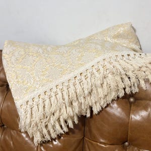 May include: A rectangular throw cushion featuring a cream and pale yellow damask pattern. The cushion is finished with a long, thick, cream-coloured fringe trim along the bottom edge, adding texture to the woven fabric surface of the cushion.