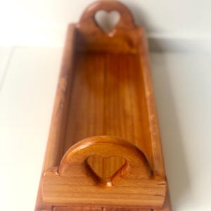May include: A rectangular wooden tray with a warm brown finish and a smooth, polished surface. The tray features two raised end handles, each containing a centred heart-shaped cutout. The base is flat with a slightly wider, bevelled edge design.