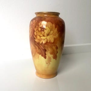 May include: A decorative vase with a floral design. The vase is painted in yellow and brown, with a large yellow flower on the front. It has a rounded shape and a narrow neck, with a gold rim. The vase is approximately 20 cm tall.