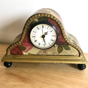 May include: A mantel clock with an arched top, featuring a floral pattern of red and white roses on the body. The clock has a gold-painted frame, a white face with black Roman numerals, ornate black hands, and four small black ball-shaped feet.