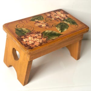 May include: A rectangular wooden step stool with a light brown finish. The top surface features a hand-painted design of pink and orange hydrangea flowers with green leaves and dark brown swirling vine accents. The side panels have heart-shaped cutouts.