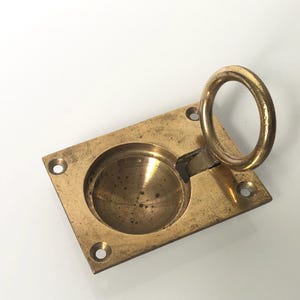 May include: A vintage brass recessed pull with a circular handle. The rectangular plate has a circular indentation and four screw holes. The handle is a ring that pivots up from the plate.