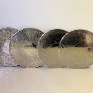 May include: Four silver decorative plates with detailed floral designs. The plates are arranged in a row, reflecting light and creating a shiny appearance. The plates have a circular shape with a central indentation. The plates are made of metal.