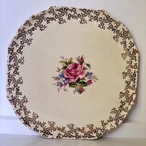 Pode incluir: Prato de servir octogonal creme com um design floral. O prato apresenta um buquê central de rosas cor-de-rosa e flores azuis. Uma borda de pequenos padrões florais dourados emoldura a borda.