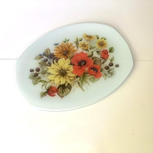 May include: A decorative glass serving tray with a floral design. The tray has a light blue background and features a bouquet of yellow, orange, and red flowers, along with green leaves and berries. The tray is oval-shaped.