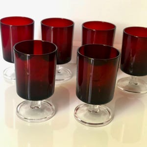 May include: Six ruby red stemmed glasses with clear bases. The glasses have a cylindrical shape and a deep red colour that fades to a darker shade at the base. The clear stems and bases provide a contrast to the rich colour of the glass.