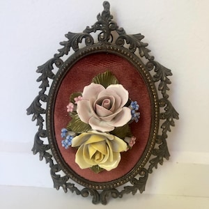 May include: An oval framed wall decor piece featuring a velvet background and a ceramic floral arrangement. The arrangement includes a pink and a yellow rose, with small blue and pink accent flowers. The frame is ornate, with a dark bronze finish.