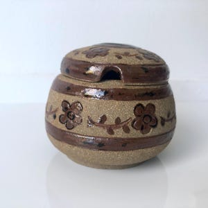 Peut inclure: Un pot en céramique beige moucheté de forme ronde avec un couvercle assorti. Le pot présente des bandes horizontales brun foncé et un motif floral peint à la main avec des fleurs et des vignes brunes. Une encoche est découpée dans le couvercle.