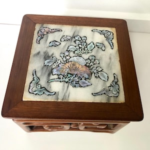 Könnte beinhalten: Kleine, quadratische Holzbox mit Marmorplatte. Der Marmor ist mit einem Blumendesign in Rosa-, Orange- und Grüntönen eingelegt. Die Box hat einen dunkelbraunen Holzrahmen und dekorative Schnitzereien an den Seiten.