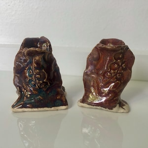 May include: Two ceramic sculptures with a textured, organic form. The pieces are glazed in shades of brown, burgundy, and teal, with a glossy finish. The bases are a light cream colour. The sculptures have a unique, artistic appearance.