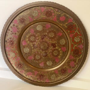 Op de afbeelding: Een ronde, antieke messing schaal met ingewikkelde bloemmotieven in roze, rood en goud. De schaal heeft een decoratieve, geschulpte rand en een centraal bloemmotief. De achtergrond is een warme, neutrale kleur.