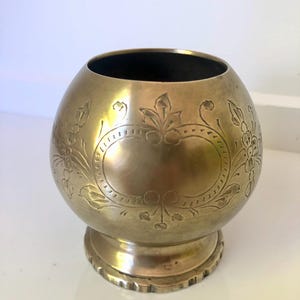 May include: A brass-colored, spherical vase with a wide opening and a pedestal base. The vase features an engraved floral design around the body, with an oval cartouche in the center. The interior is dark.
