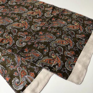May include: A dark olive green scarf with a repeating paisley pattern in orange, white, and yellow. The scarf has a light beige border and is made of a soft, flowing fabric. The scarf is folded and displayed on a white surface.